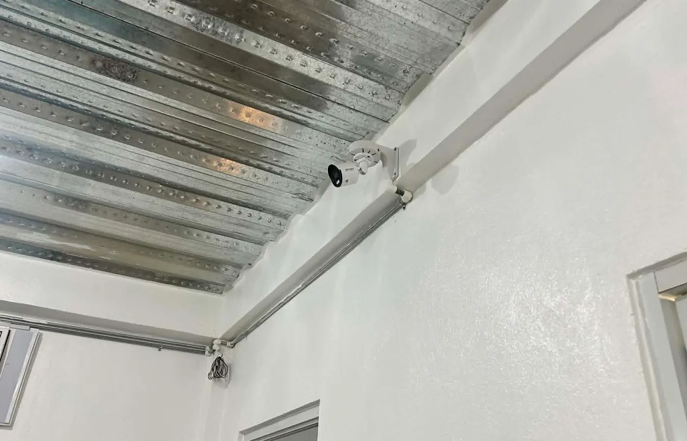 A CCTV camera mounted at a ceiling corner, wired through steel roof decking, overlooking a room below.