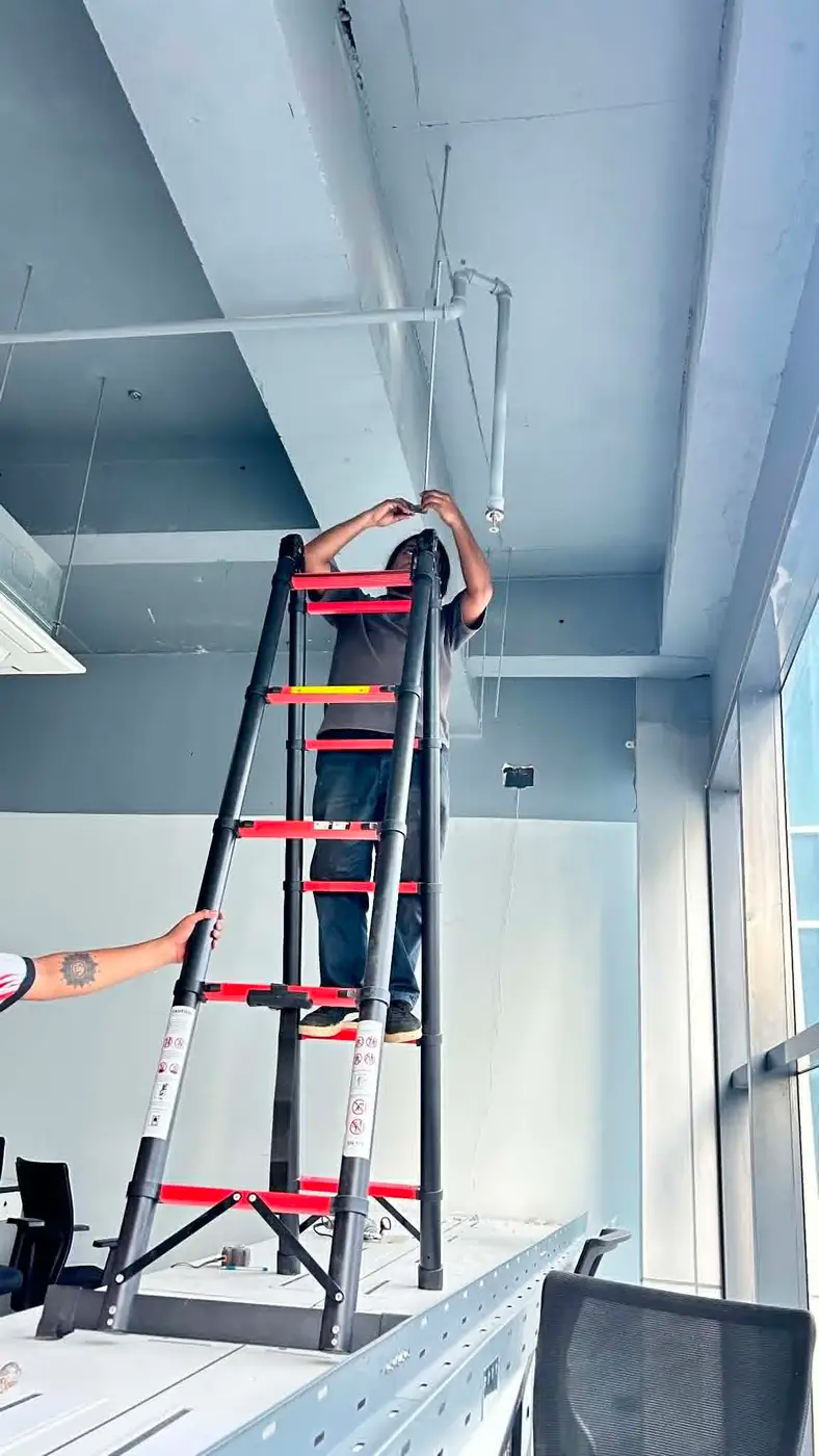Technician on a tall ladder reaching into the ceiling void to route network cabling, assisted by a colleague holding the ladder.