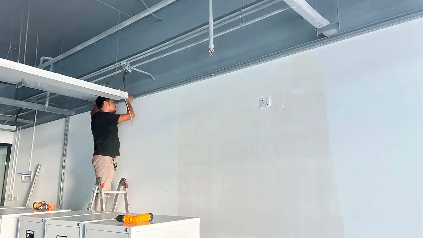 Technician on a ladder installing a cable trunking channel along an office wall.