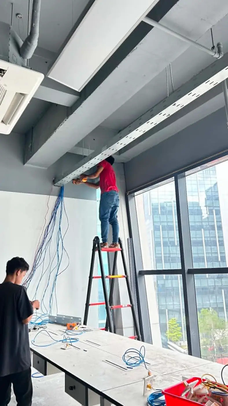Technician installing ceiling cable trunking near a window overlooking Mactan Newtown, with a colleague terminating cables below.