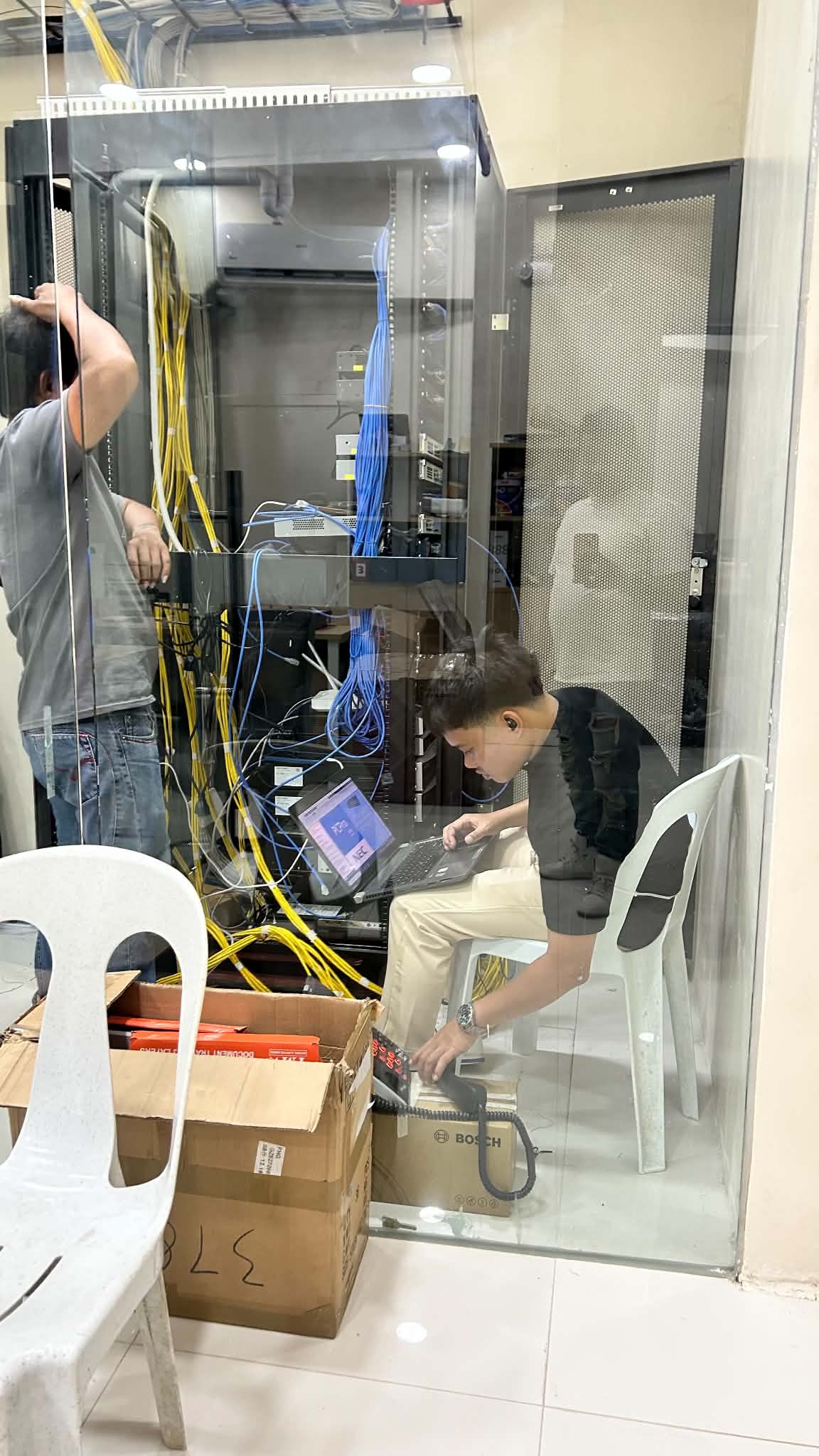 Technician seated in the server room testing terminated cable runs on a laptop while a colleague works on cable management above.