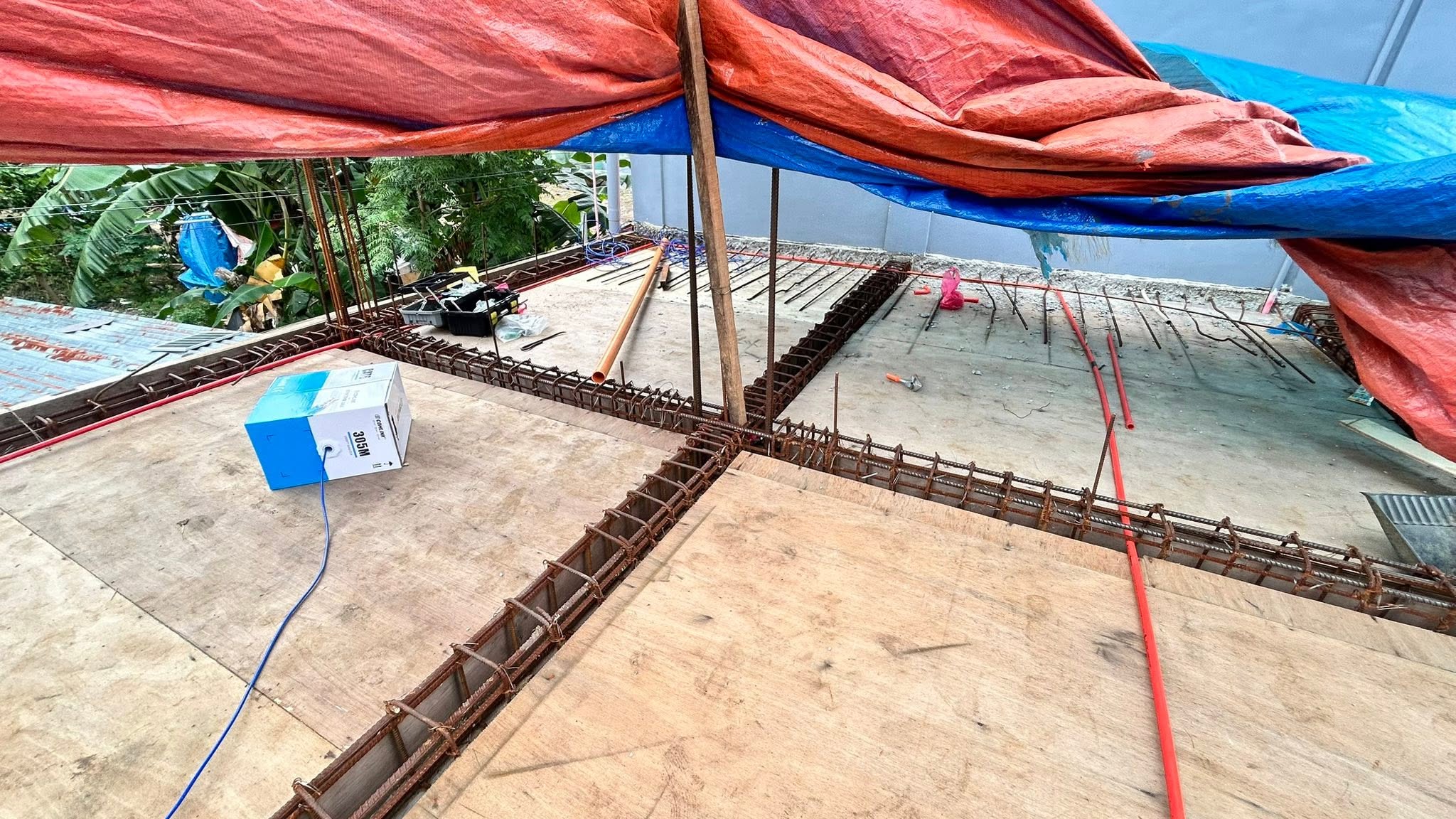 A boxed cable spool feeding blue cable along a red conduit run laid across a plywood roof deck under a tarp canopy.