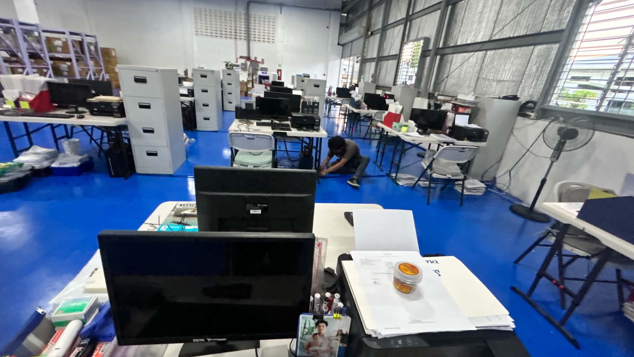 Inside the warehouse office area with tables, monitors, filing cabinets, and active workstations set up across the floor.