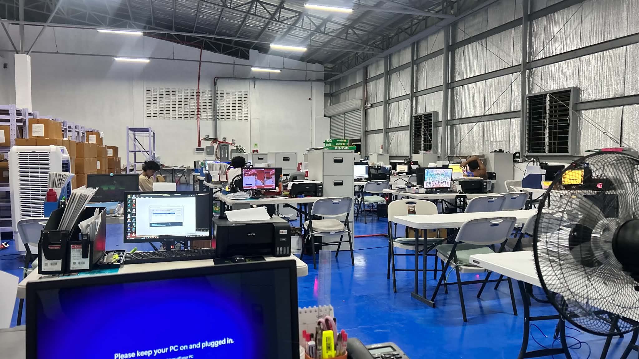 A wide warehouse office view showing desks, storage racks, and open circulation space for staff and inventory movement.