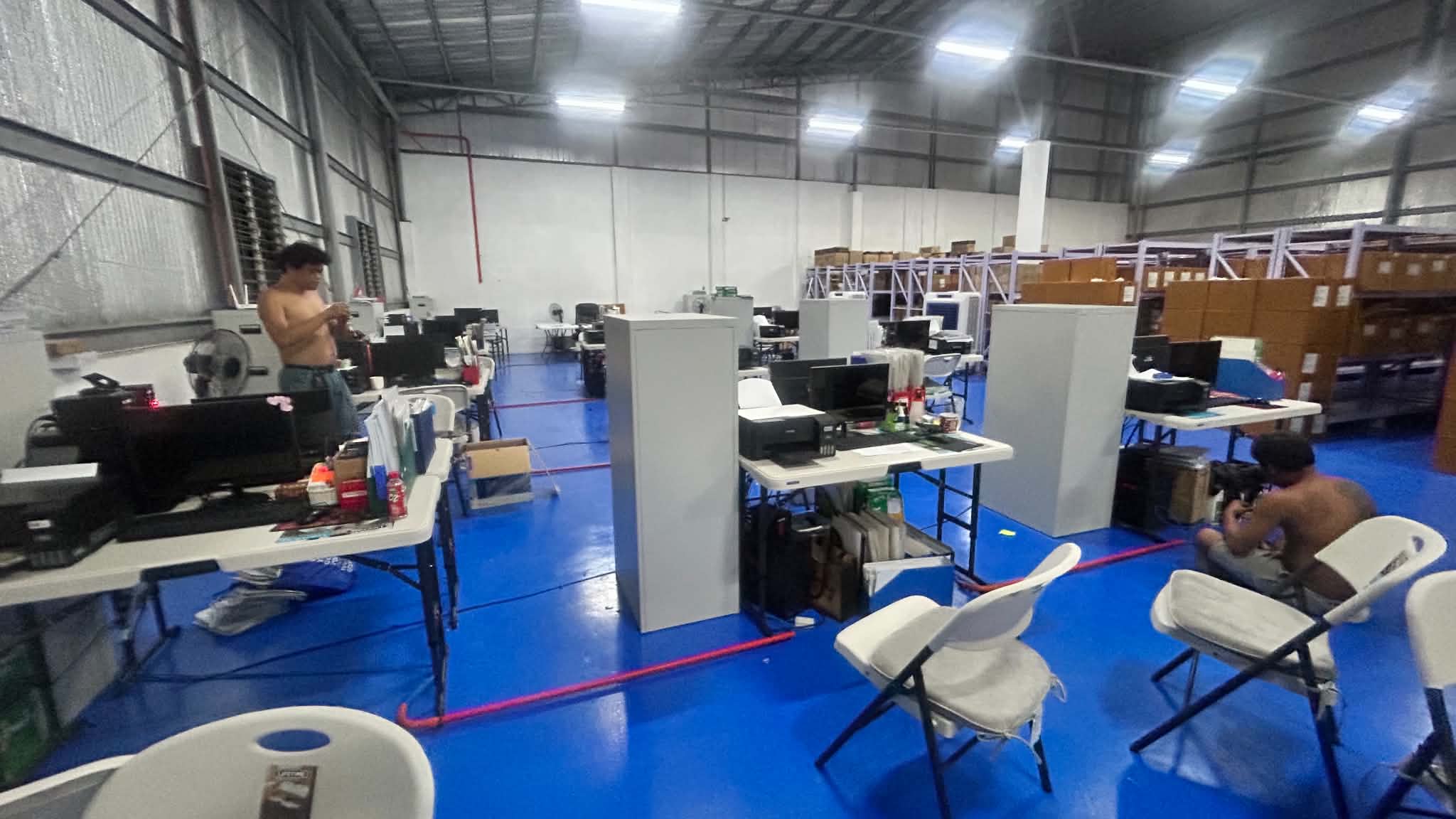 Warehouse office setup with multiple desks and computers positioned under high-bay lighting beside boxed inventory racks.