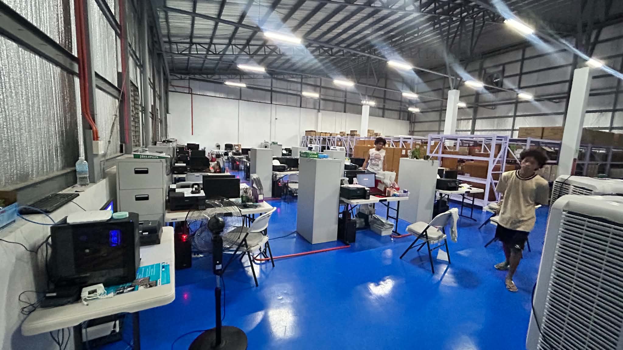 Another wide angle of the warehouse office showing desks, monitors, cabinets, and storage shelves in a live working environment.
