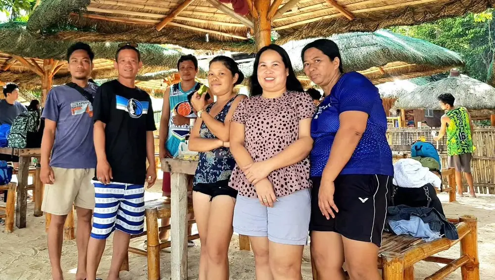 Six teammates posing together under a nipa hut roof at the beach.