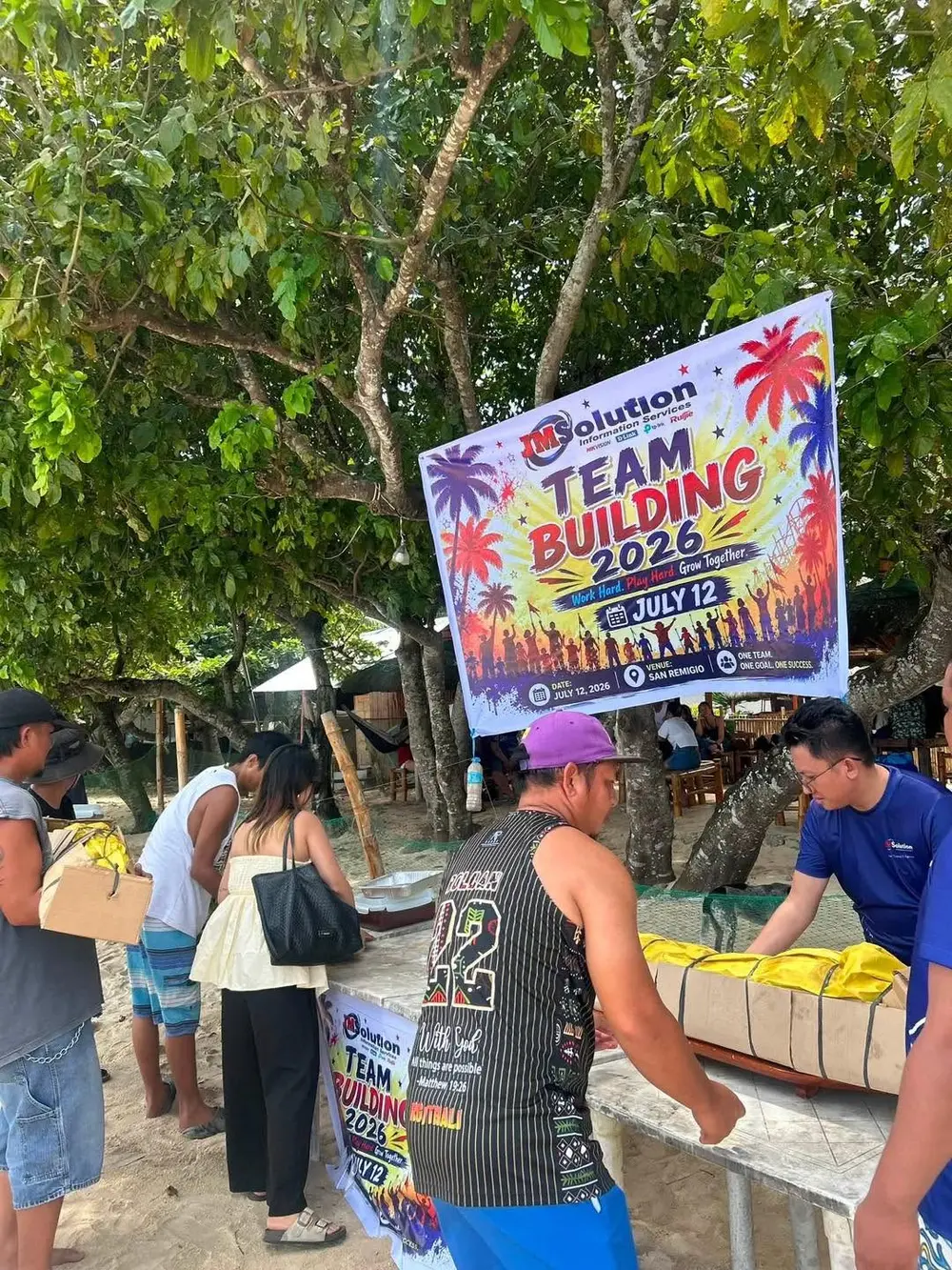 Teammates and family gathered near the food tables and event banner under the trees at the beach.